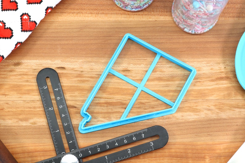 May include: A light blue plastic cookie cutter shaped like a house with a grid pattern. The cutter is on a wooden surface with a ruler and other baking supplies.
