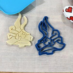 May include: Two cookie cutters, one blue and one beige, shaped like a dragon's head. The cutters are on a white surface.