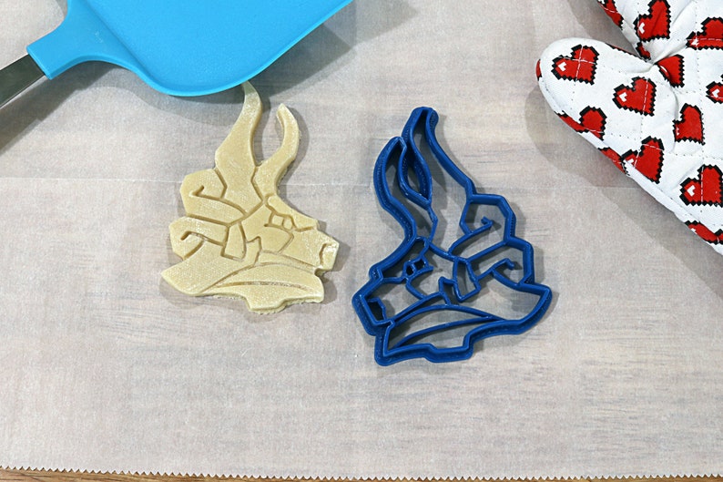 May include: Two cookie cutters, one blue and one beige, shaped like a dragon's head. The cutters are on a white surface.