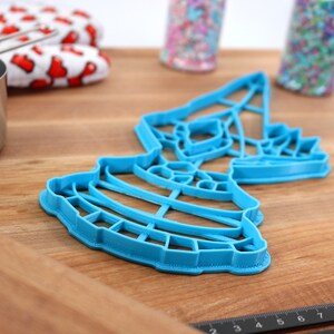 May include: A blue plastic cookie cutter shaped like a tiered cake with a star on top. The cutter is on a wooden surface.