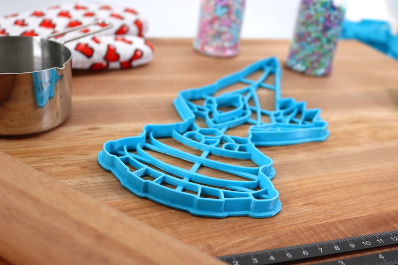 May include: A blue plastic cookie cutter shaped like a tiered cake with a star on top. The cutter is on a wooden surface.