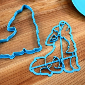 May include: Two blue plastic cookie cutters, one shaped like a wolf and the other shaped like a tree, on a wooden surface.