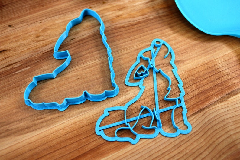 May include: Two blue plastic cookie cutters, one shaped like a wolf and the other shaped like a tree, on a wooden surface.