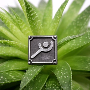 Peut inclure: Une &eacute;pingle en m&eacute;tal argent&eacute; avec une image stylis&eacute;e d'une personne tenant une loupe. L'&eacute;pingle est de forme carr&eacute;e et a un fond noir. L'&eacute;pingle est pos&eacute;e sur une plante succulente verte.