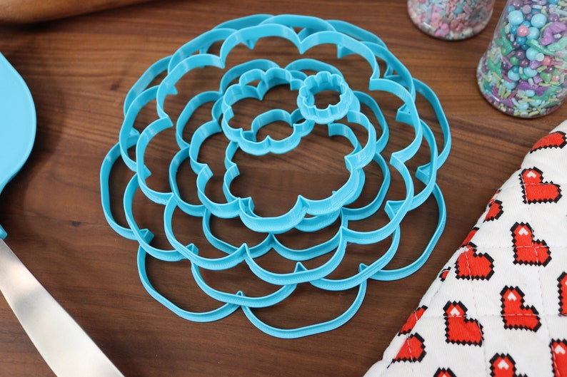 May include: A set of six blue plastic cookie cutters shaped like a flower. The cutters are nested inside each other, with the largest cutter at the bottom and the smallest cutter at the top.