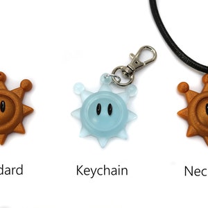 May include: Three versions of a gold-colored sun-shaped pendant with black eyes. The pendant is available as a standard charm, a keychain, and a necklace.