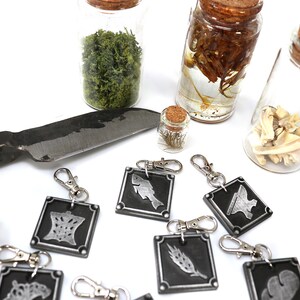 May include: A set of eight silver keychains with different symbols on black backgrounds. The keychains are arranged on a white surface with a knife, a small bottle of dried flowers, and three glass jars containing various items.
