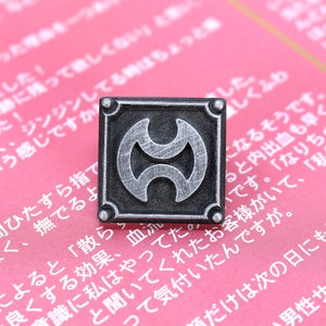 May include: A silver metal pin with a black background and a stylized symbol of two overlapping crescent moons. The pin is on a pink surface with Japanese text.