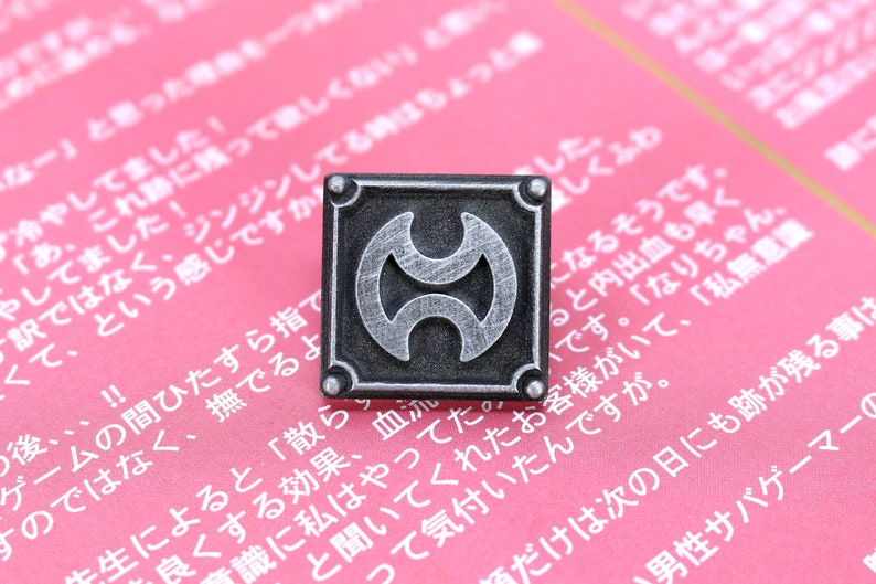 May include: A silver metal pin with a black background and a stylized symbol of two overlapping crescent moons. The pin is on a pink surface with Japanese text.