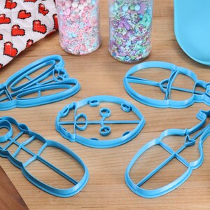 May include: Five blue plastic cookie cutters shaped like a ladybug, a beetle, a dragonfly, a stag beetle, and a caterpillar. The cutters are on a wooden surface.