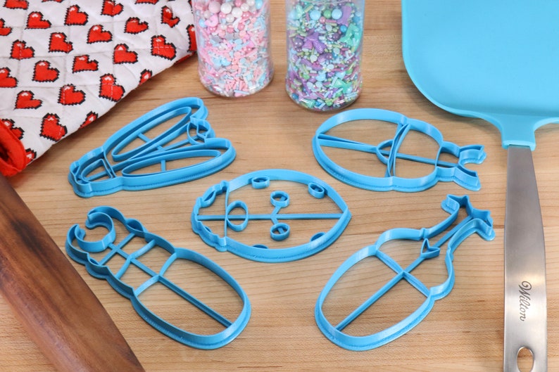 May include: Five blue plastic cookie cutters shaped like a ladybug, a beetle, a dragonfly, a stag beetle, and a caterpillar. The cutters are on a wooden surface.
