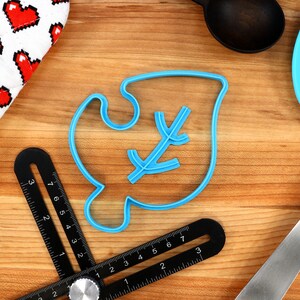 May include: Blue plastic cookie cutter shaped like a leaf with a small branch inside. The cutter is on a wooden surface with a ruler and a spatula.