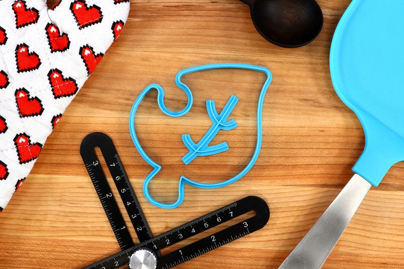 May include: Blue plastic cookie cutter shaped like a leaf with a small branch inside. The cutter is on a wooden surface with a ruler and a spatula.