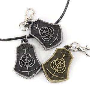 May include: Two silver and one gold pendant necklaces with a black leather cord. Each pendant features a shield-shaped design with a stylized symbol of three overlapping circles and a vertical line.