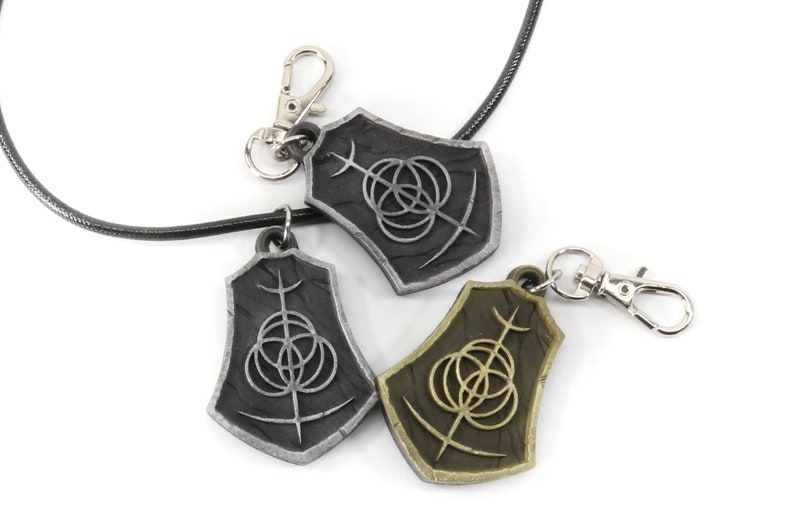May include: Two silver and one gold pendant necklaces with a black leather cord. Each pendant features a shield-shaped design with a stylized symbol of three overlapping circles and a vertical line.