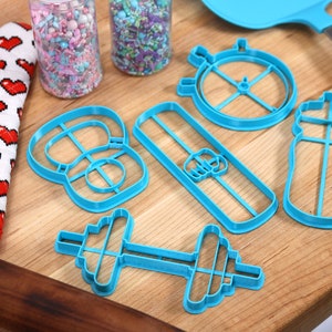 May include: A set of blue plastic cookie cutters shaped like a dumbbell, a water bottle, a kettlebell, a protein bar, and a stopwatch. The cutters are on a wooden surface.