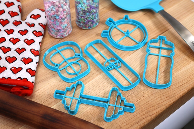 May include: A set of blue plastic cookie cutters shaped like a dumbbell, a water bottle, a kettlebell, a protein bar, and a stopwatch. The cutters are on a wooden surface.