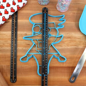 May include: A blue plastic cookie cutter shaped like a cartoon character with a large hat and a long nose. The cookie cutter is on a wooden surface with a ruler and a spatula.