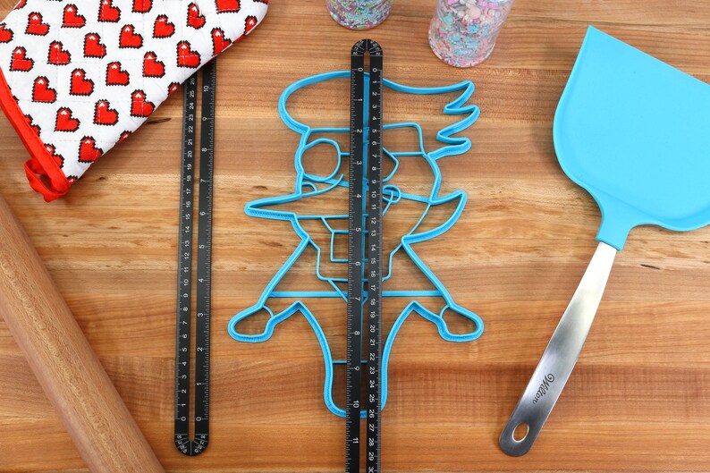 May include: A blue plastic cookie cutter shaped like a cartoon character with a large hat and a long nose. The cookie cutter is on a wooden surface with a ruler and a spatula.
