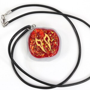 May include: A red and gold pendant with a black cord necklace. The pendant is a square shape with a white border and a red and gold design inside.