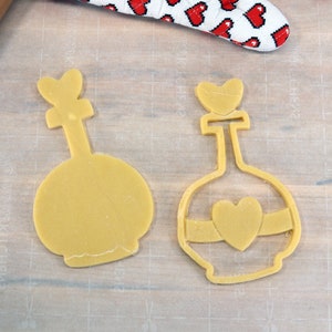 May include: Two yellow cookie cutters shaped like bottles with heart-shaped tops. The bottle on the right has a heart-shaped design on the front.