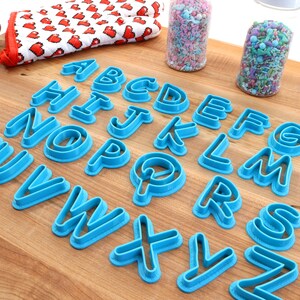Op de afbeelding: Een set blauwe plastic alfabetkoekjesvormen op een houten oppervlak. De vormen zijn alfabetisch gerangschikt.
