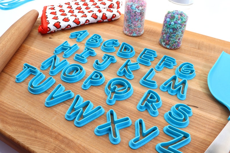 Op de afbeelding: Een set blauwe plastic alfabetkoekjesvormen op een houten oppervlak. De vormen zijn alfabetisch gerangschikt.