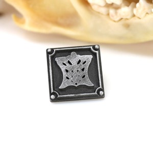 May include: A silver and black metal square pin with a decorative design in the center.