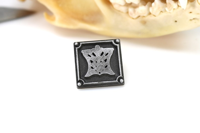 May include: A silver and black metal square pin with a decorative design in the center.