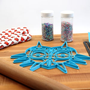 May include: A blue plastic cookie cutter shaped like the Majora's Mask from the Legend of Zelda video game. The cutter is on a wooden cutting board with a rolling pin, a pair of oven mitts, and two jars of sprinkles.