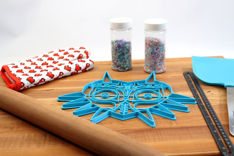May include: A blue plastic cookie cutter shaped like the Majora's Mask from the Legend of Zelda video game. The cutter is on a wooden cutting board with a rolling pin, a pair of oven mitts, and two jars of sprinkles.