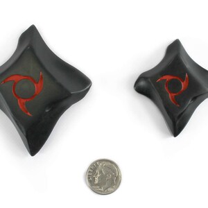 K&ouml;nnte beinhalten: Zwei schwarze, rautenf&ouml;rmige Objekte mit rotem Spiraldesign, m&ouml;glicherweise dekorativ oder funktional. Eine M&uuml;nze dient zum Gr&ouml;&szlig;envergleich. Die Objekte haben eine leicht gekr&uuml;mmte, strukturierte Oberfl&auml;che und sind vor wei&szlig;em Hintergrund platziert.