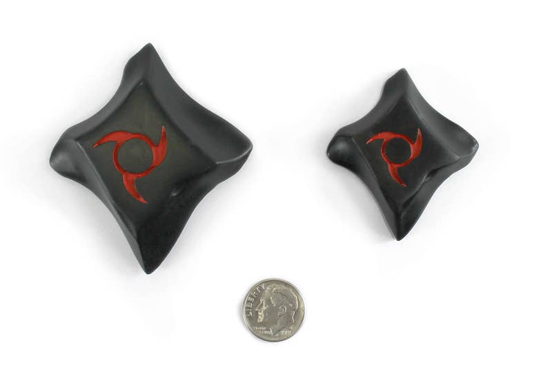 K&ouml;nnte beinhalten: Zwei schwarze, rautenf&ouml;rmige Objekte mit rotem Spiraldesign, m&ouml;glicherweise dekorativ oder funktional. Eine M&uuml;nze dient zum Gr&ouml;&szlig;envergleich. Die Objekte haben eine leicht gekr&uuml;mmte, strukturierte Oberfl&auml;che und sind vor wei&szlig;em Hintergrund platziert.