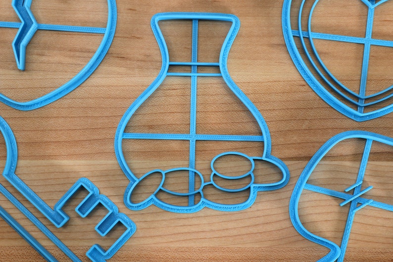 May include: A set of five blue plastic cookie cutters in various shapes, including a vase, a key, a heart, and two leaves. The cutters are on a wooden surface.