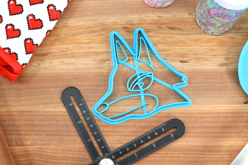 May include: A blue plastic cookie cutter shaped like a dog's head. The cutter is on a wooden surface.