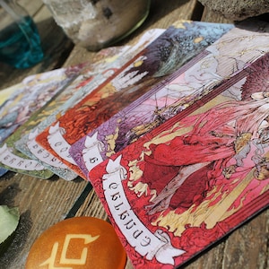 May include: A spread of tarot cards on a wooden table. The cards are colorful and feature intricate illustrations. The top card shows a woman with wings, holding a scale and a sword. The card is labeled "Judgment".