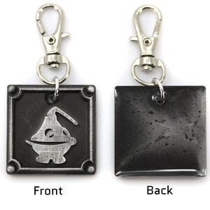 May include: A silver metal keychain with a black square charm. The front of the charm features a stylized silver design of a creature with a single eye. The back of the charm is plain.