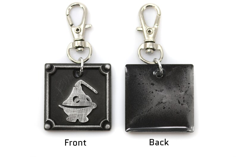 May include: A silver metal keychain with a black square charm. The front of the charm features a stylized silver design of a creature with a single eye. The back of the charm is plain.