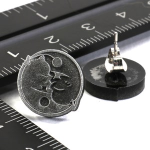 May include: A pair of silver stud earrings featuring a black and white design of a bat and a crescent moon. The earrings are shown next to a ruler.