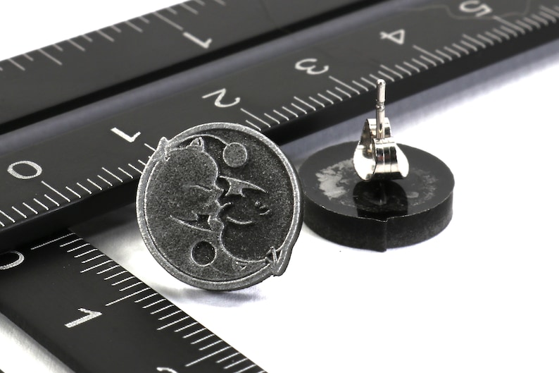 May include: A pair of silver stud earrings featuring a black and white design of a bat and a crescent moon. The earrings are shown next to a ruler.