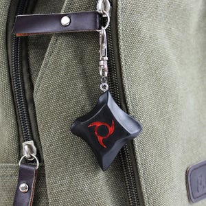 K&ouml;nnte beinhalten: Schwarzer und roter Anh&auml;nger mit einzigartigem Design, befestigt an einem silberfarbenen Verschluss. Der Anh&auml;nger h&auml;ngt an einem gr&uuml;nen Canvas-Rucksack mit braunen Lederakzenten. Das Design des Anh&auml;ngers ist eine rote Spirale.