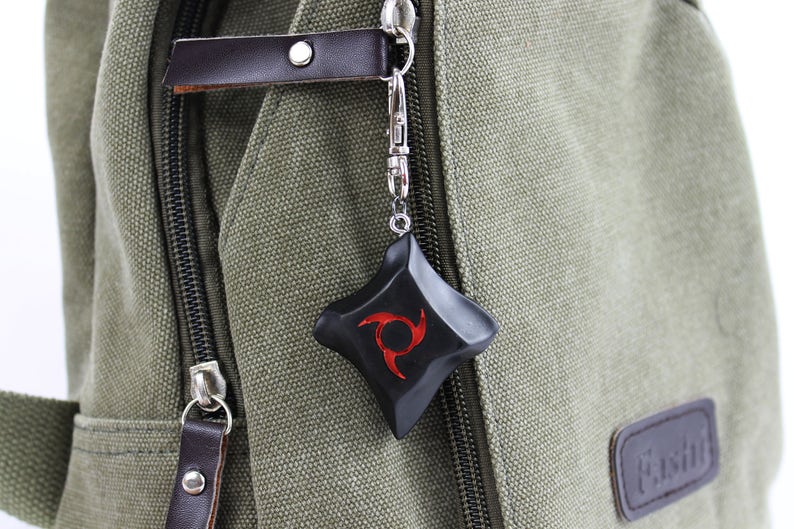 K&ouml;nnte beinhalten: Schwarzer und roter Anh&auml;nger mit einzigartigem Design, befestigt an einem silberfarbenen Verschluss. Der Anh&auml;nger h&auml;ngt an einem gr&uuml;nen Canvas-Rucksack mit braunen Lederakzenten. Das Design des Anh&auml;ngers ist eine rote Spirale.