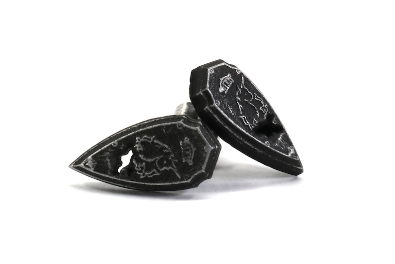 May include: A pair of black metal cufflinks shaped like shields with a silver design of a mythical creature.