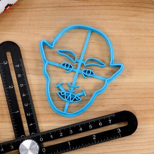 May include: A blue plastic cookie cutter shaped like a face with a mischievous expression. The cutter has a wide mouth with sharp teeth, raised eyebrows, and large eyes.