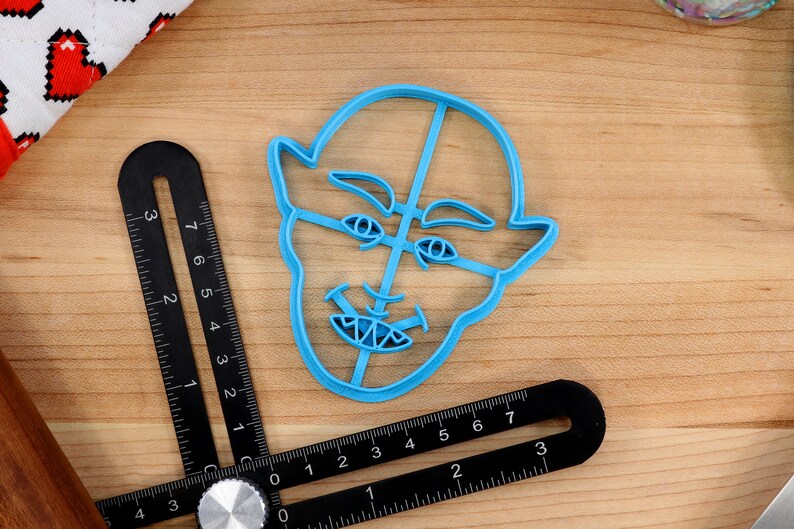 May include: A blue plastic cookie cutter shaped like a face with a mischievous expression. The cutter has a wide mouth with sharp teeth, raised eyebrows, and large eyes.