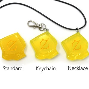 May include: Three yellow pendants with an orange design, labeled "Standard", "Keychain", and "Necklace". The "Keychain" version has a silver clasp. The "Necklace" version is on a black cord. All are on a white background.
