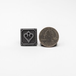 May include: A small, square, silver-colored metal charm with a stylized heart design. The charm is about the size of a US quarter.