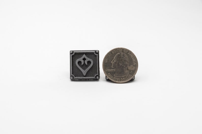 May include: A small, square, silver-colored metal charm with a stylized heart design. The charm is about the size of a US quarter.