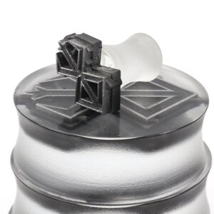 May include: A silver metal container with a black rubber stamp on top. The stamp has a geometric design.