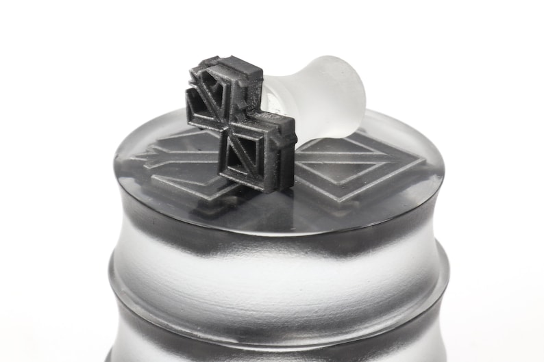 May include: A silver metal container with a black rubber stamp on top. The stamp has a geometric design.
